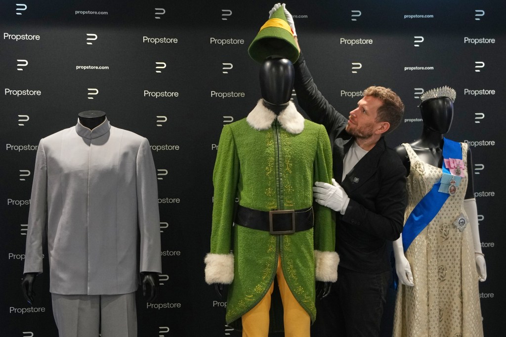 A person adjusting Buddy the Elf's costume, displayed next to a light gray suit and a queen's dress.