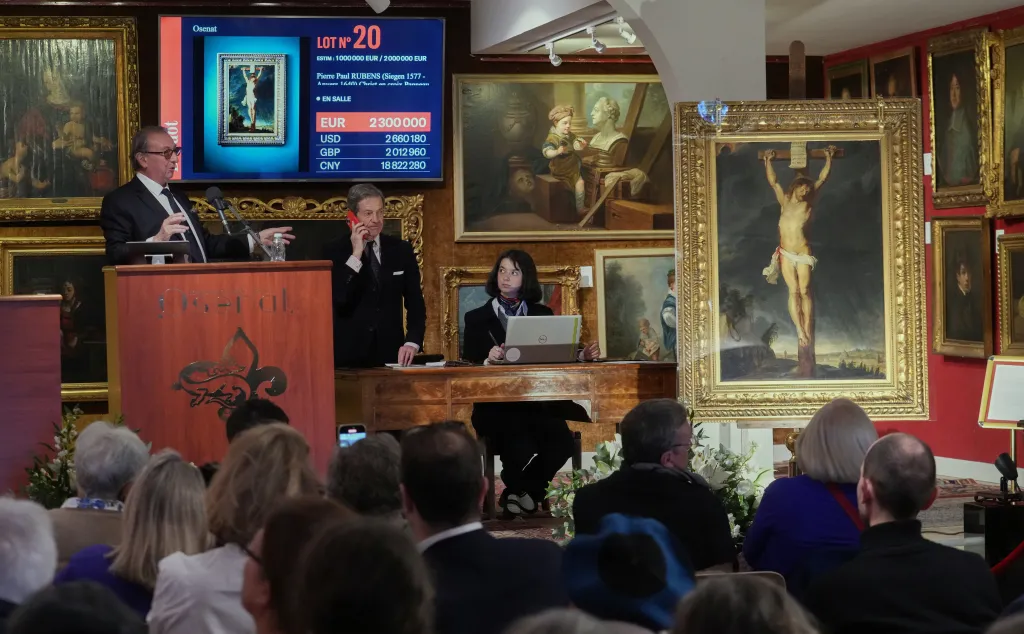 An auction room with a man at a podium, another man on a phone, and a woman at a laptop, with a large screen displaying 