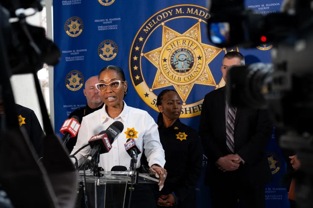 Sheriff Melody M. Maddox speaking at a podium with microphones, flanked by other officers, against a backdrop of the DeKalb County Sheriff's Office logo.