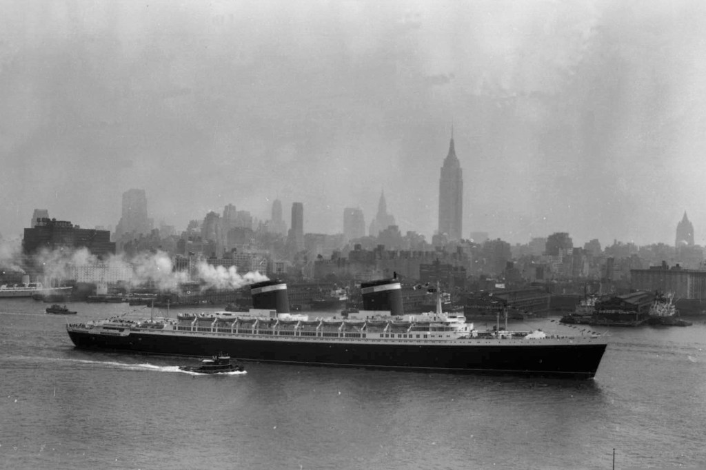 A large ship with two smokestacks moving through water with a city skyline in the background.