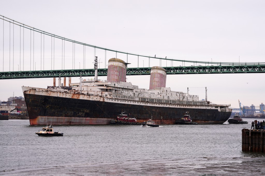 The historic ship S.S. United States, a large rusted ocean liner, being guided by tugboats under a green suspension bridge.
