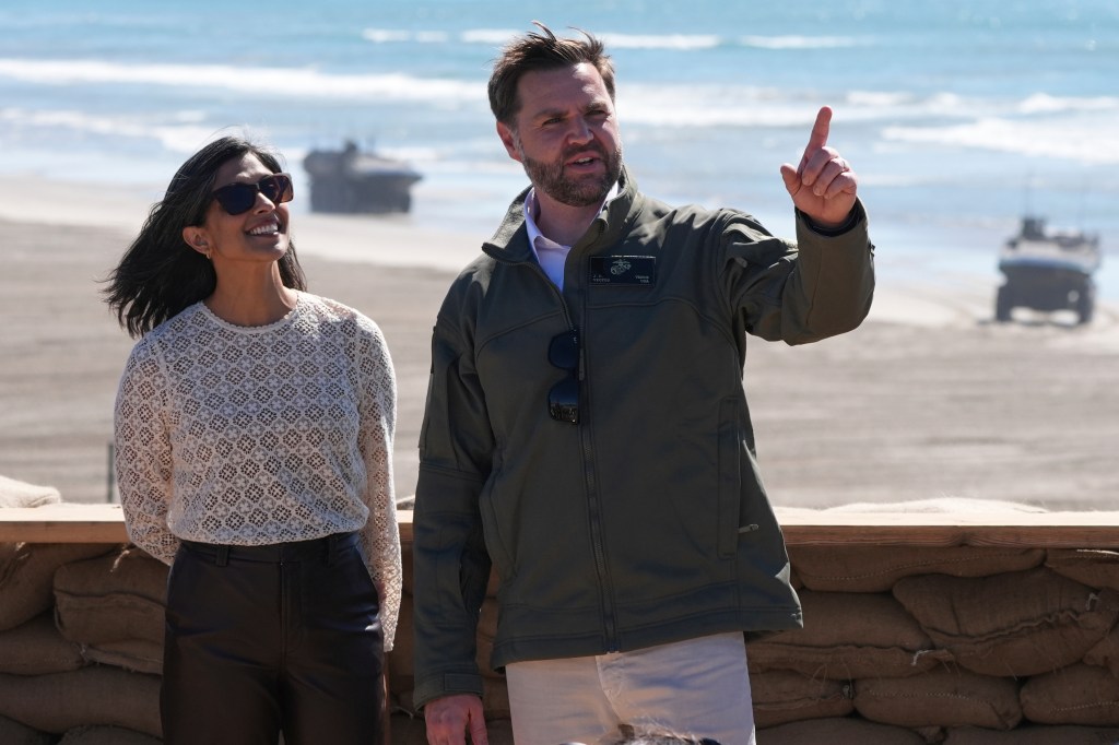 JD Vance and a woman standing in front of a beach with military vehicles in the background.