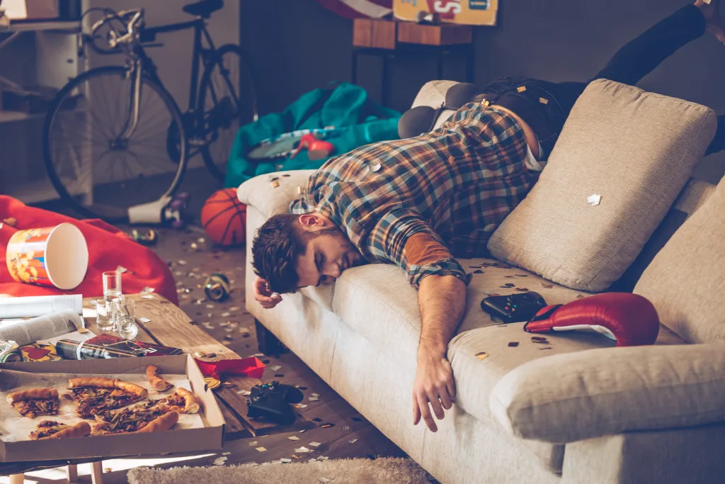A messy room after a party, with a young man asleep on a couch.