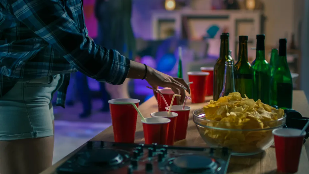 A person in a plaid shirt reaching for a drink in a red cup at a party, with snacks, empty bottles, and a DJ mixer on a table.