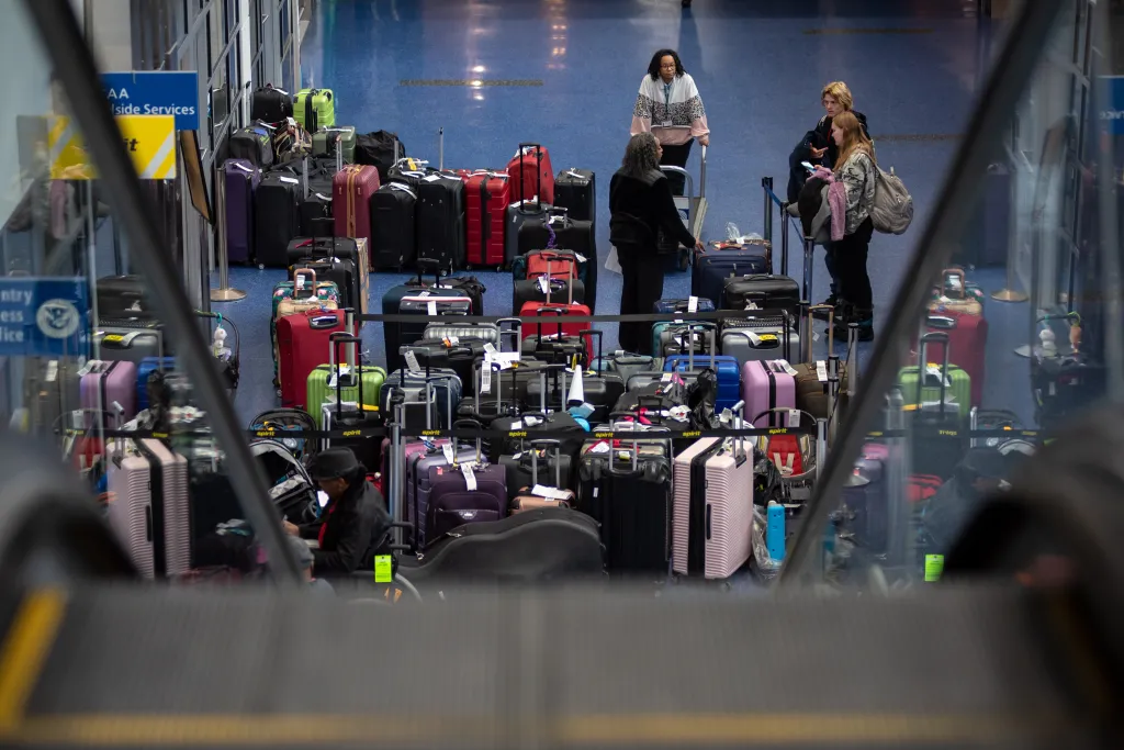 Passengers and a large number of suitcases are visible from an escalator looking down at a baggage claim area.