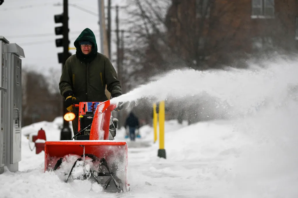A man operating a snowblower on a snowy sidewalk, with snow flying out of the snowblower's chute.