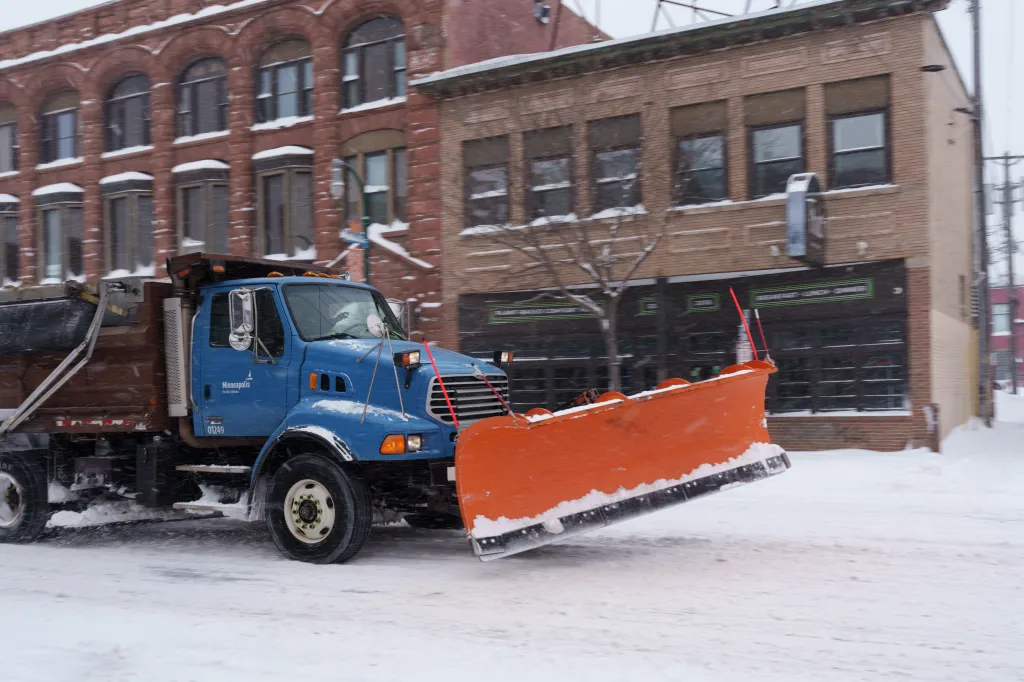 A blue snow plow truck with an orange plow clearing snow on a city street.