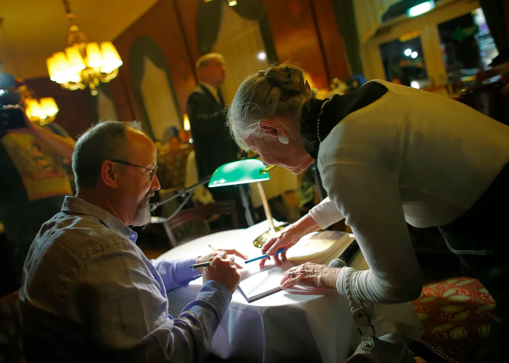 A man in glasses signing a book at a table with an older woman looking over his shoulder.