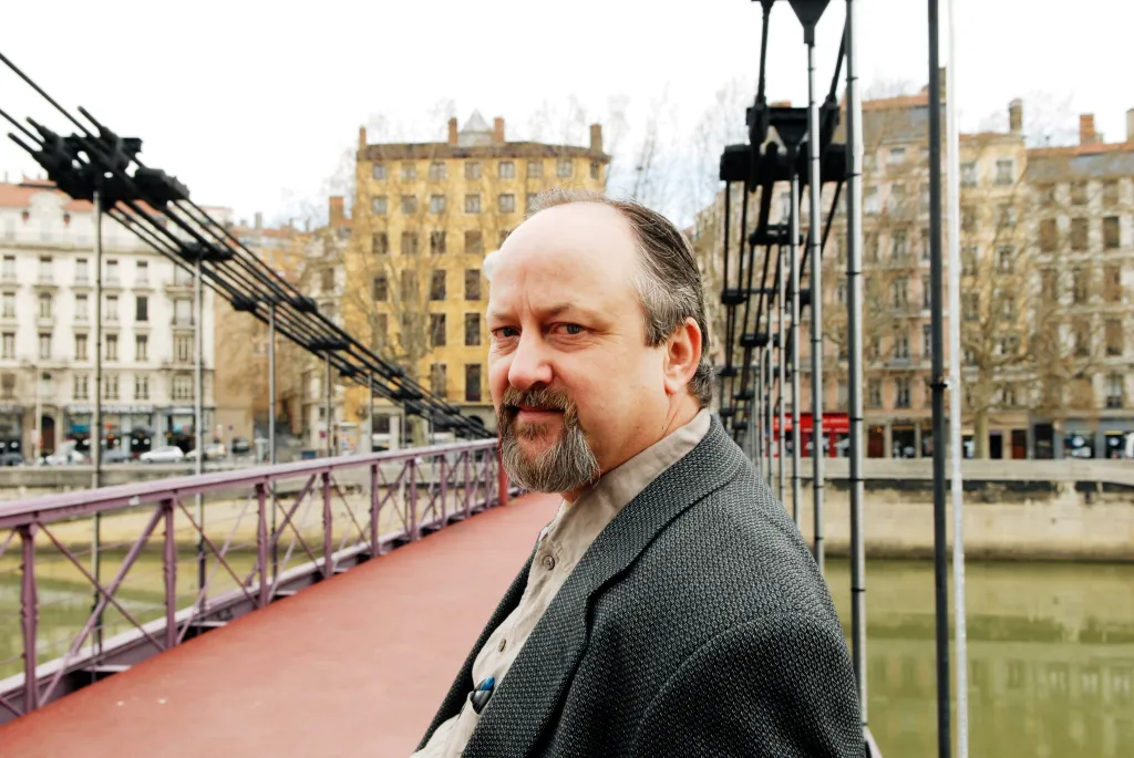 A man with a goatee and business casual attire stands on a bridge with buildings in the background.