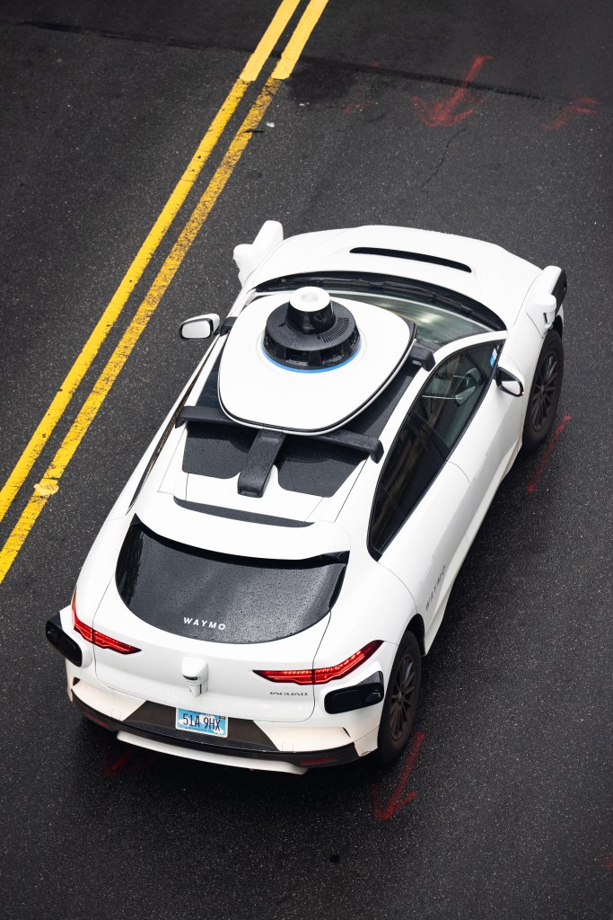 A white Waymo self-driving car on an asphalt road with double yellow lines.