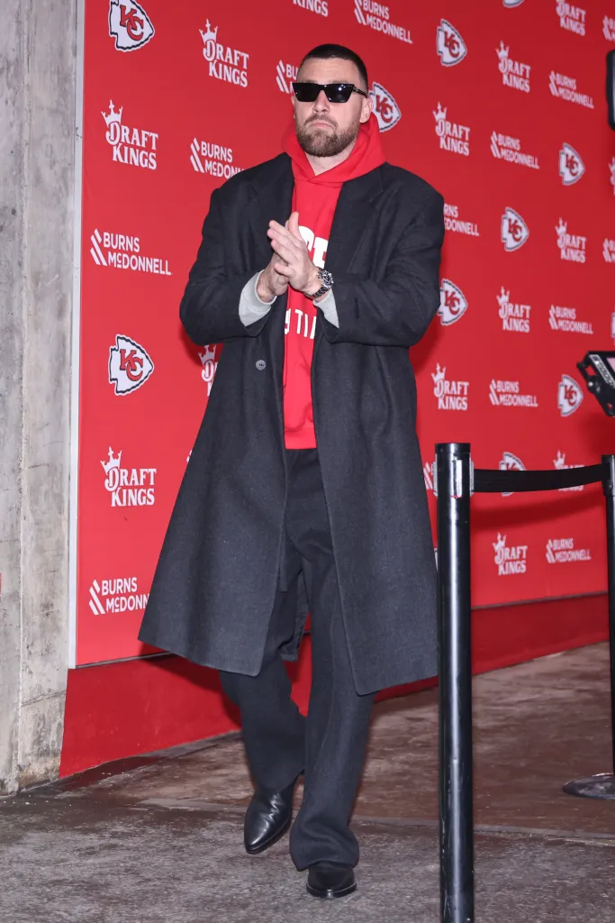 Travis Kelce in sunglasses, red hoodie, and long dark coat, clapping his hands in front of a red backdrop with 