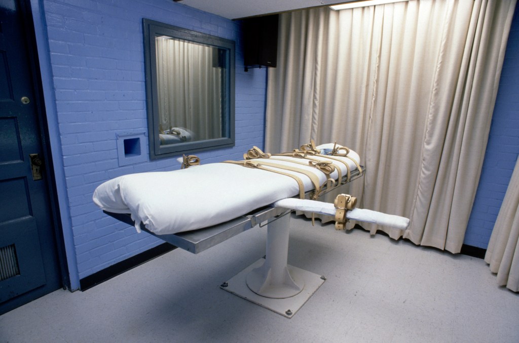 A padded execution gurney with restraints in a blue-walled room, with a mirrored window on the left and curtains on the right.