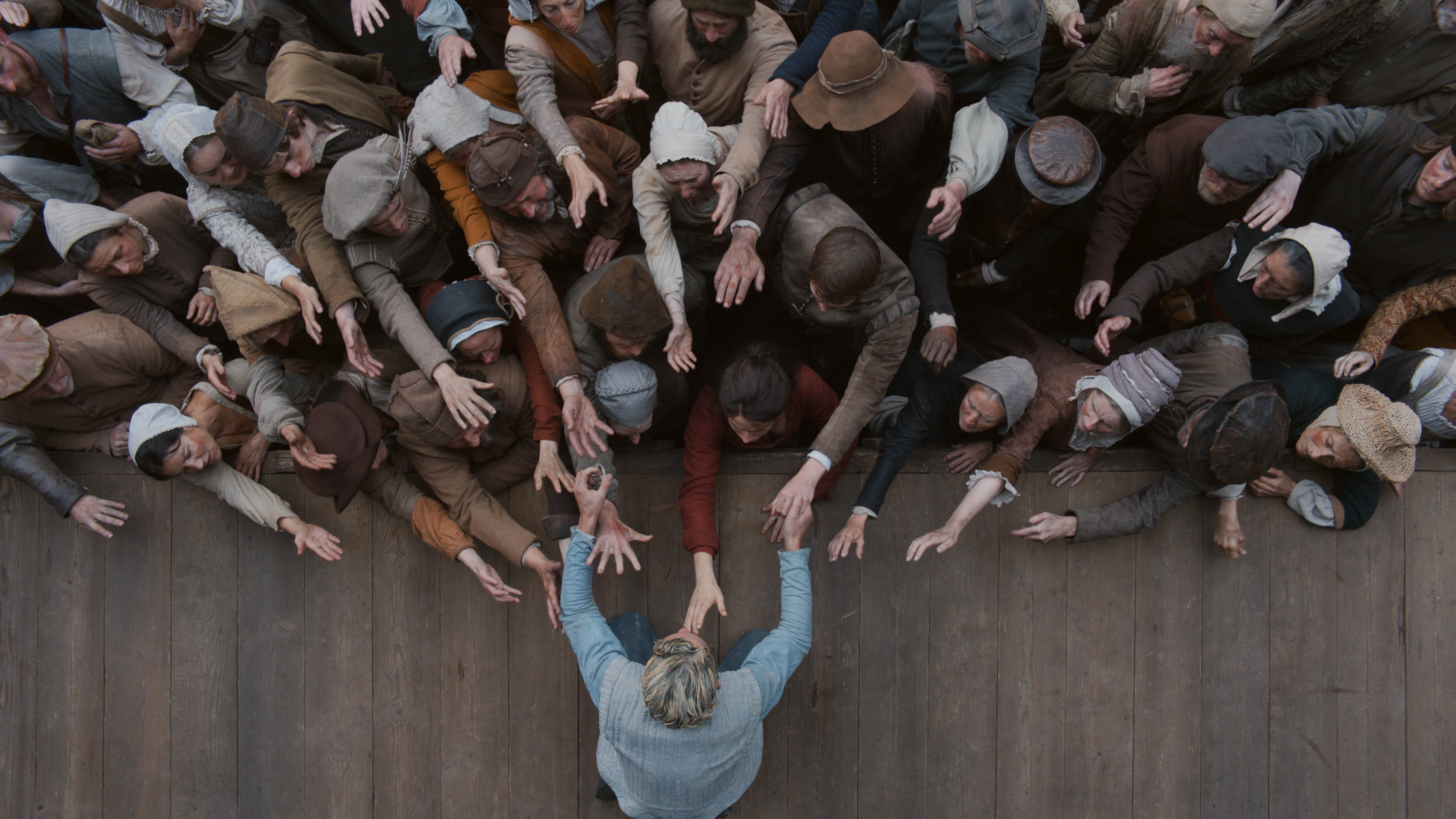 Seen from above, a blond man in blue reaches off the edge of a stage to a crowded audience, which is reaching back towards him. Seen from above, a blond man in blue reaches off the edge of a stage to a crowded audience, which is reaching back towards him.