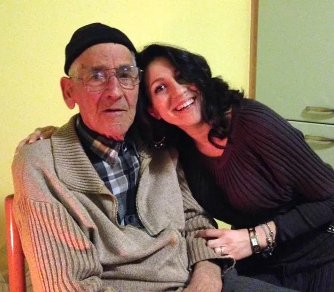 A smiling woman leans on an older man wearing a knit cap and glasses.