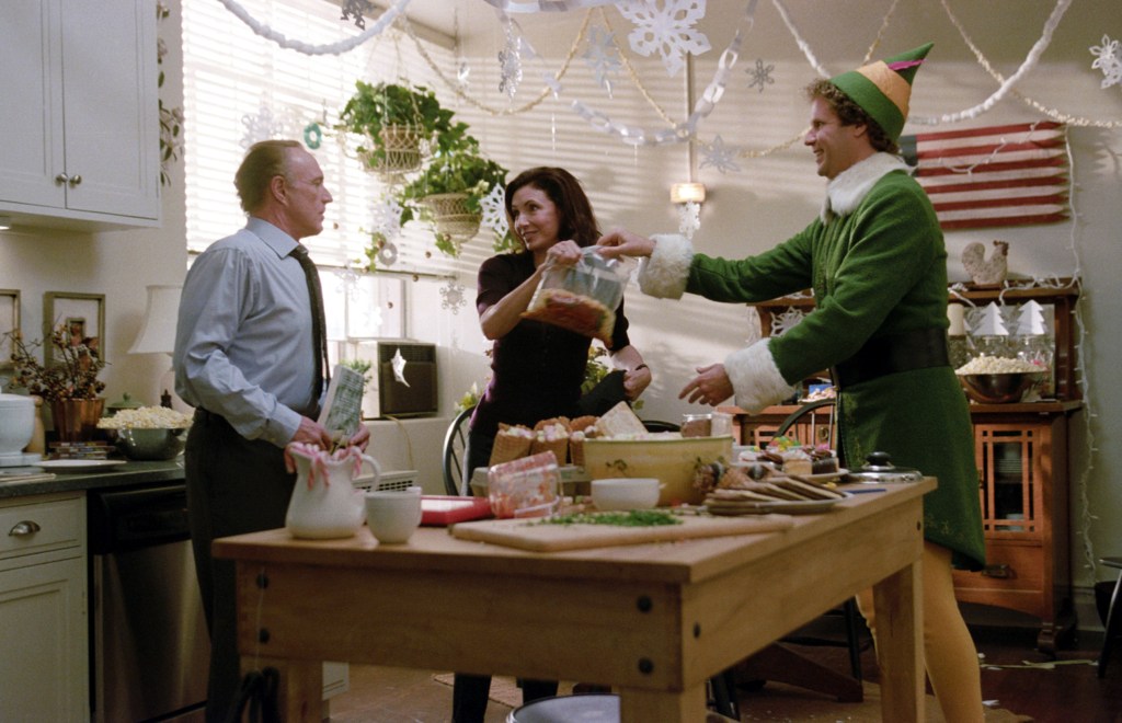 Buddy the Elf, a man in a green elf costume, smiles as he receives a bag of snacks from a woman in a decorated kitchen, while another man looks on.