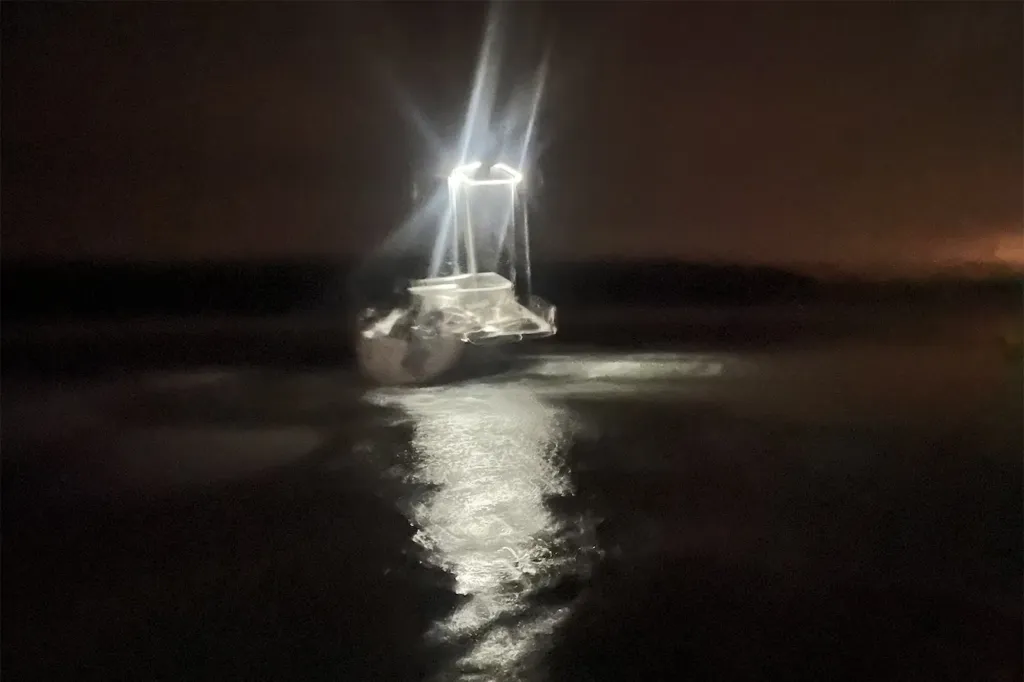 A boat on the water at night, illuminated by its own bright lights, creating a reflection on the water.
