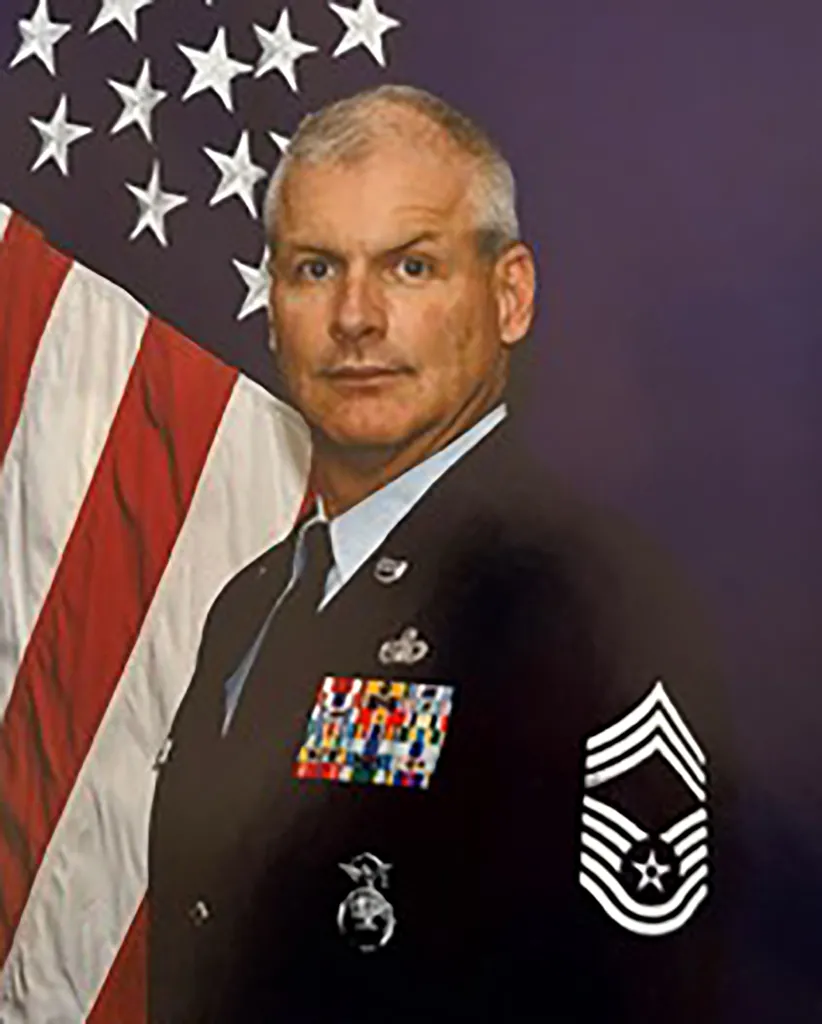 A man in a military uniform with medals and an American flag behind him.