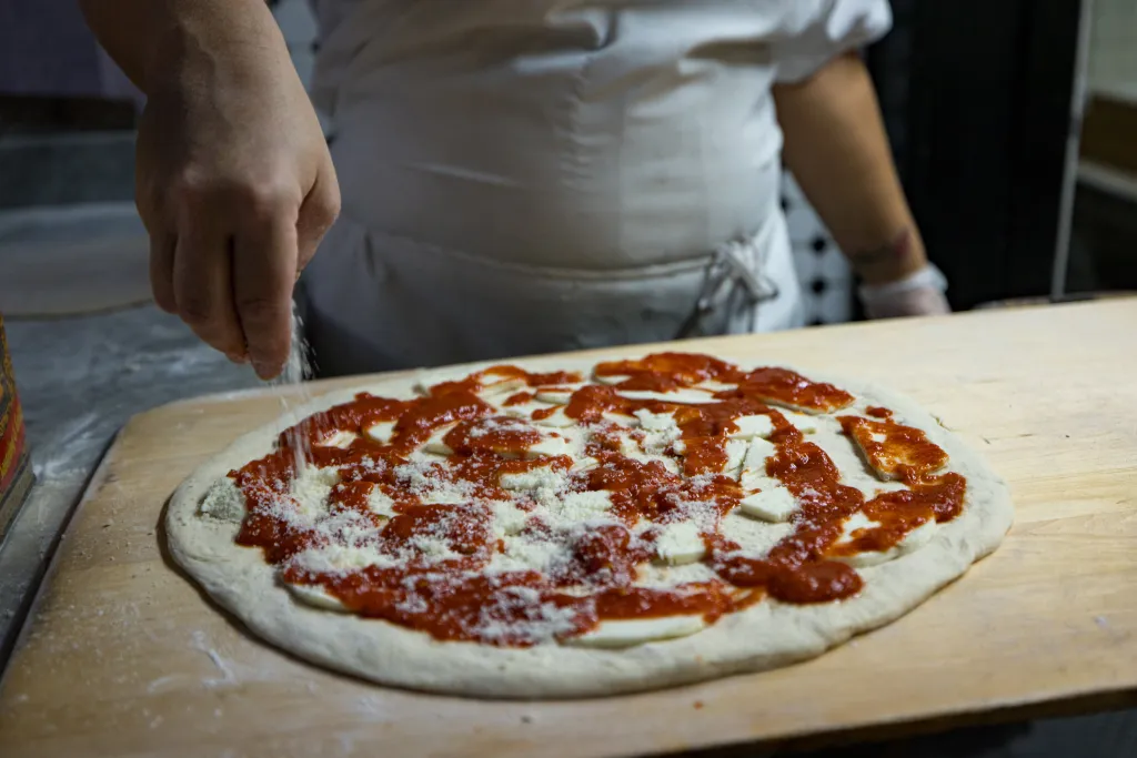 A cook's hand sprinkling cheese onto a pizza with tomato sauce and mozzarella cheese on a wooden peel.