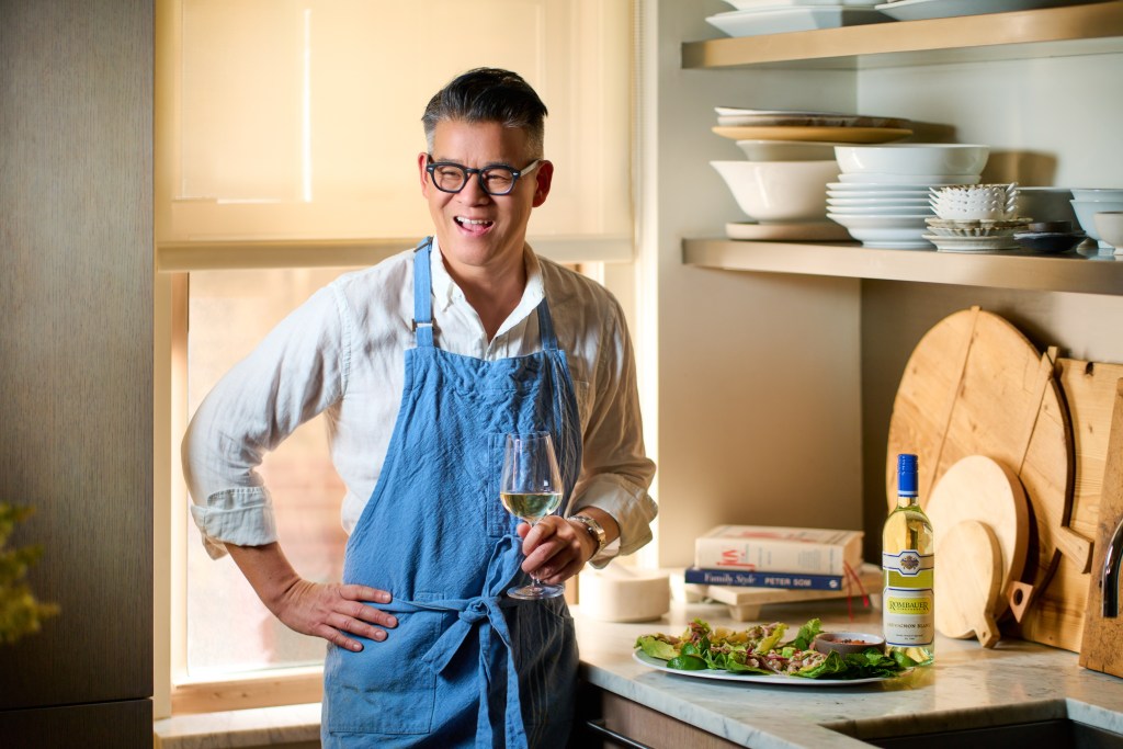Peter Som holding a glass of white wine and standing next to Mahi-Mahi Larb and a bottle of Rombauer Vineyards Sauvignon Blanc.