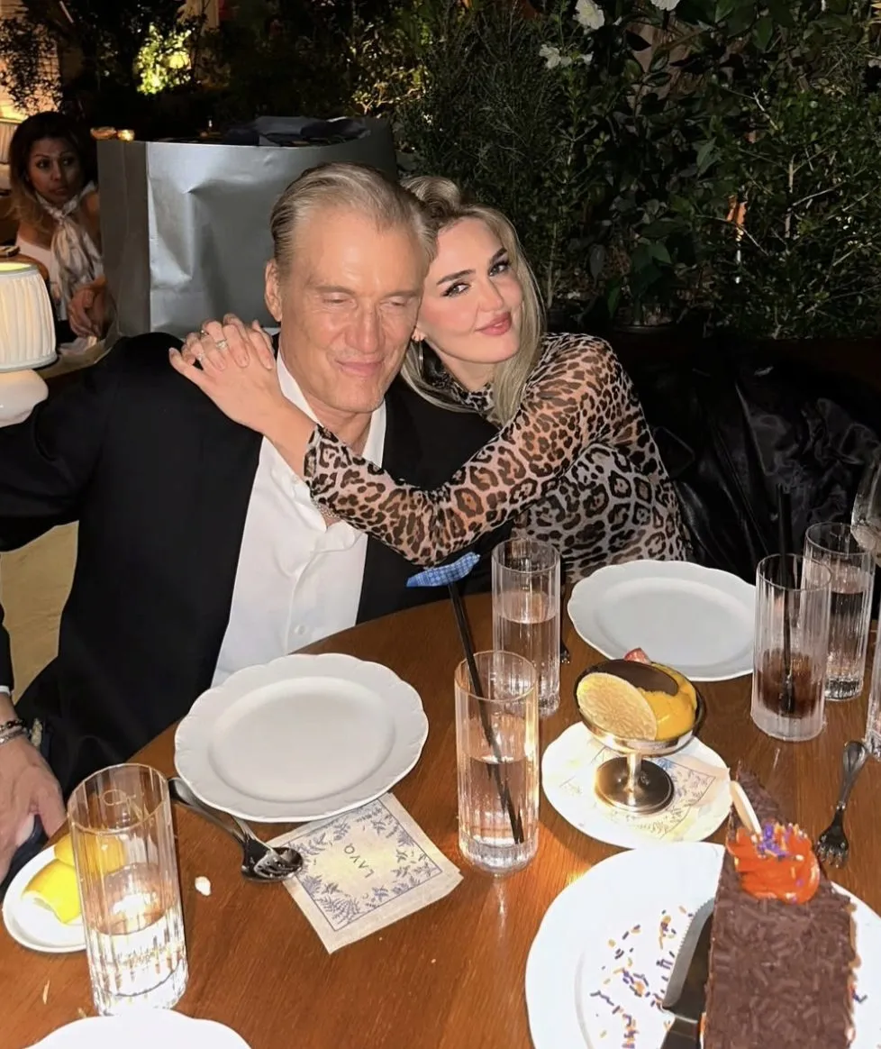 A man in a suit jacket and a woman in a leopard print dress pose together at a restaurant table with dessert.