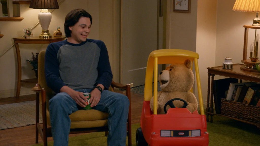 Ted, a talking teddy bear, sits in a children's car smiling at John (Max Burkholder), who sits in a chair and smiles back