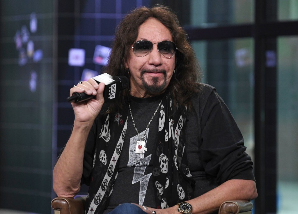 Ace Frehley holding a microphone and wearing sunglasses, a skull-patterned scarf, and a necklace with an Ace of Hearts pendant.
