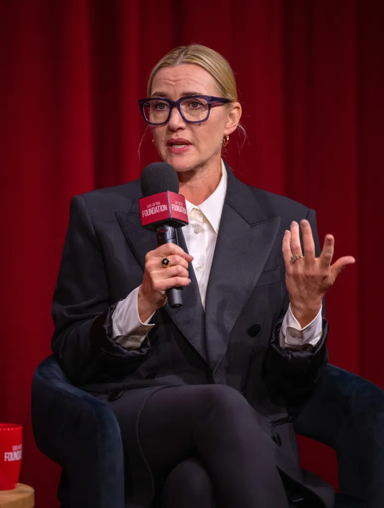 Kate Winslet, wearing dark glasses and a suit, speaks into a microphone.