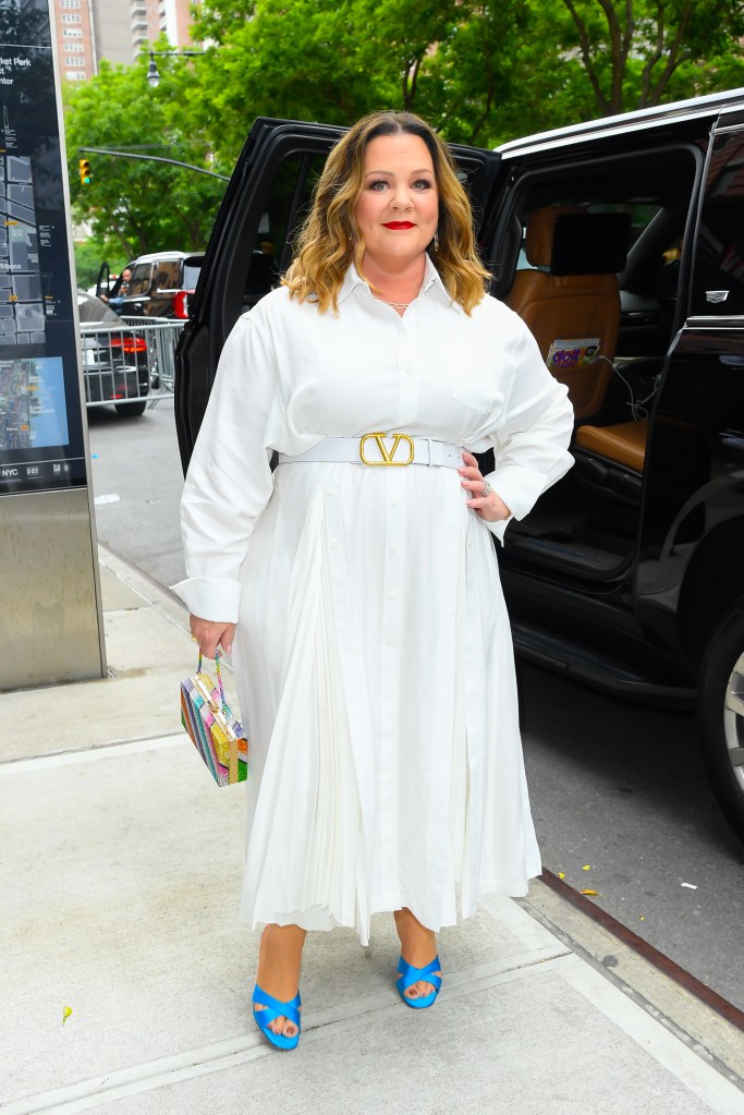 Melissa McCarthy in a white shirt dress with a white belt, blue shoes, and a colorful clutch, next to an open car door.