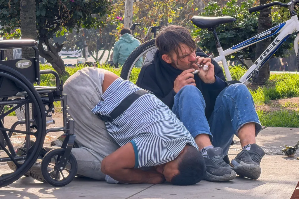 A person smoking from a pipe next to a person lying on the ground, with a wheelchair and bicycle nearby.