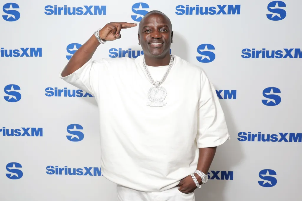 Akon smiling and pointing at his forehead in front of a SiriusXM backdrop.