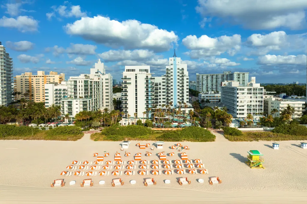 Aerial view of a beach in Miami with many beach chairs and umbrellas, with hotels and tall buildings in the background.