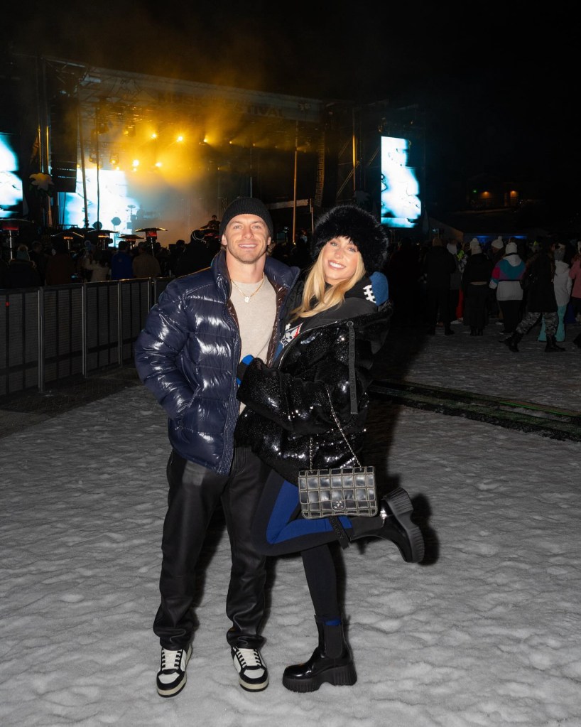 Alix Earle and Braxton Berrios smiling, wearing winter clothing in front of a stage at night.