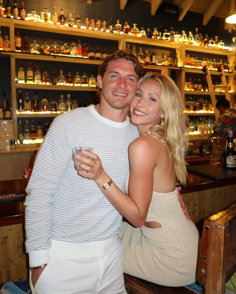 Alix Earle and Braxton Berrios smiling together at a bar with bottles on shelves in the background.