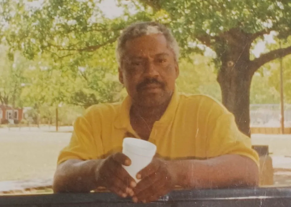 Almond Gene Little, a man with a mustache and goatee, wearing a yellow shirt and holding a white cup, sitting outdoors with trees in the background.