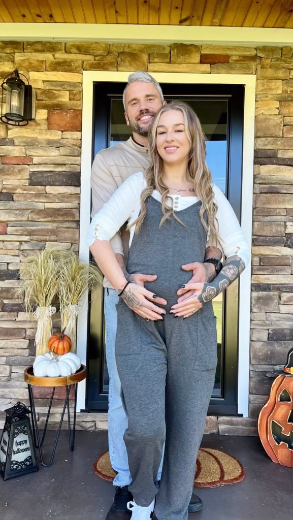 A pregnant woman in gray overalls and a man with gray hair smile, as he holds her baby bump.