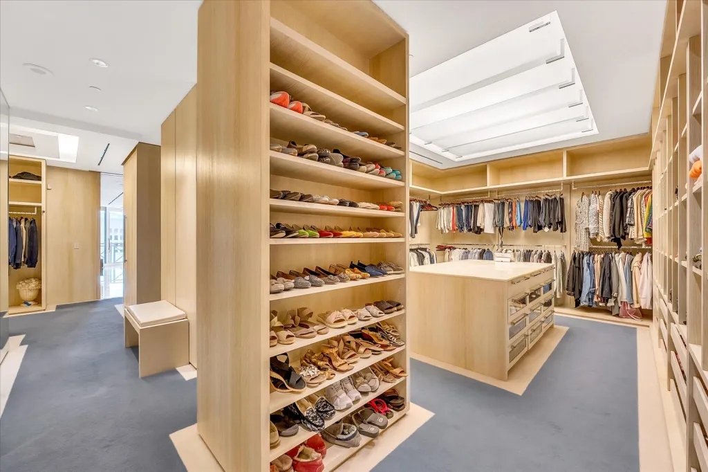 A large walk-in closet with light wood cabinetry, blue carpet, shelves filled with shoes, and clothes hanging on racks.