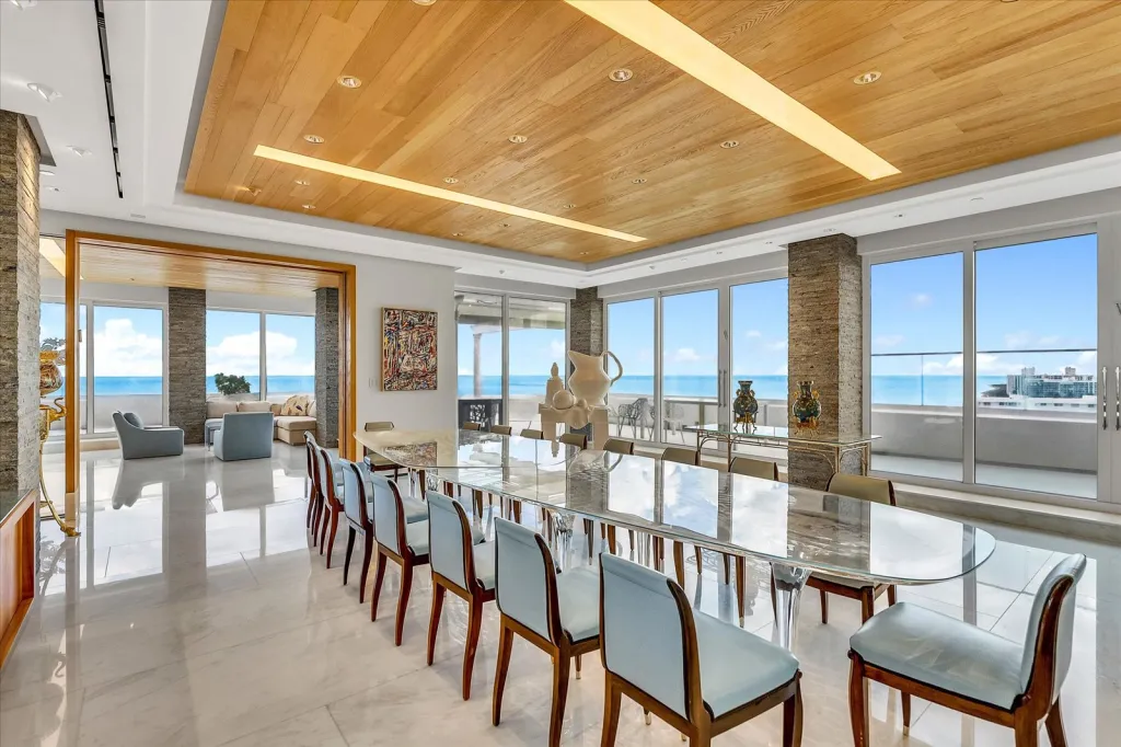 Penthouse dining room with a long glass table, light blue chairs, and floor-to-ceiling windows showing an ocean view.