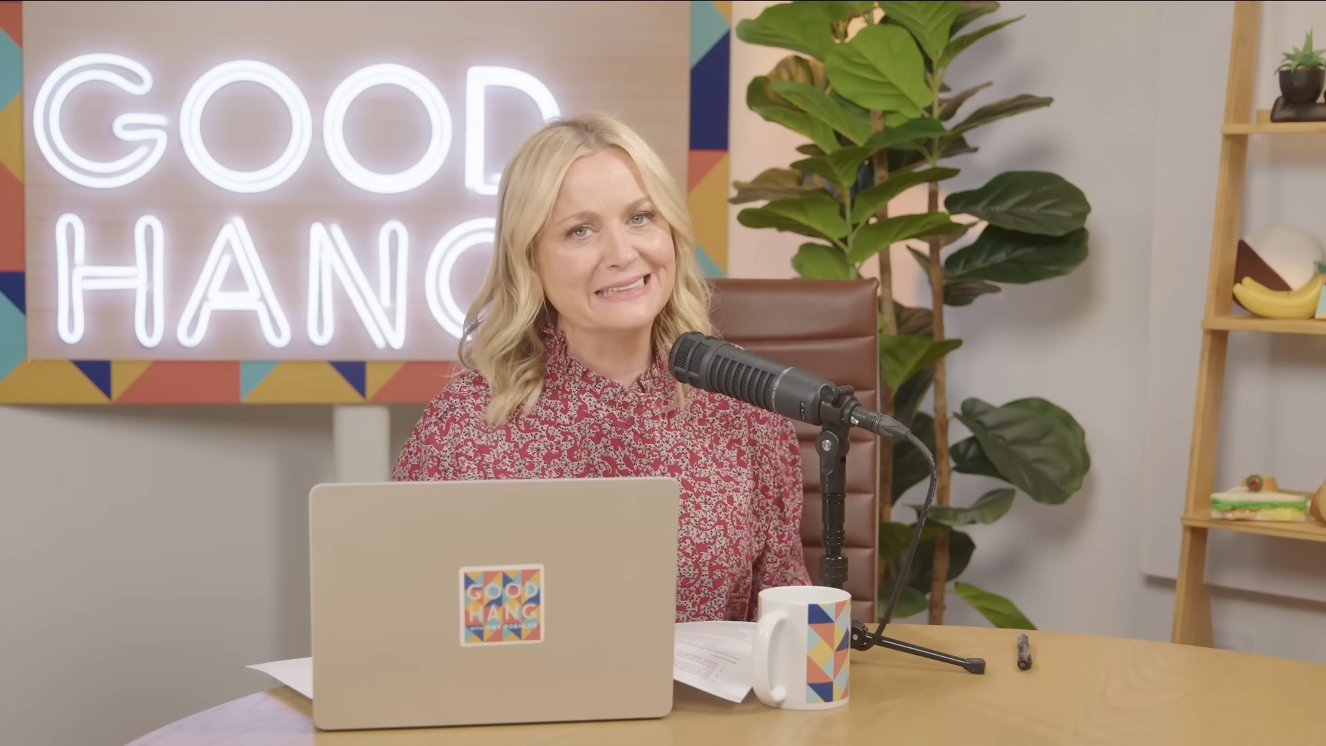 Amy Poehler in a red floral shirt, sitting at a desk with a laptop and a microphone.