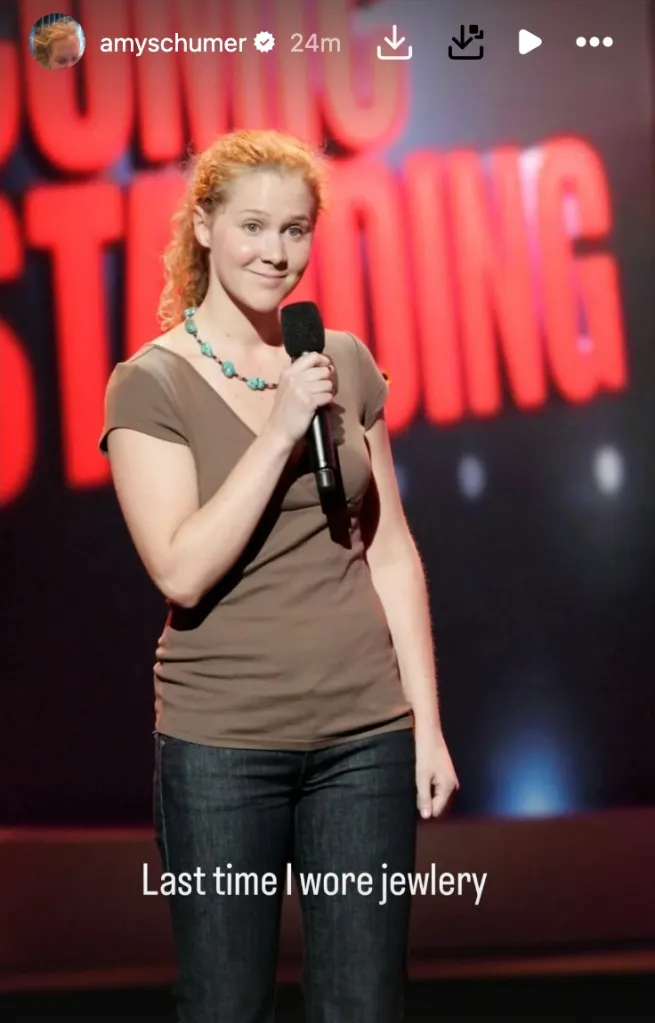 Amy Schumer on stage, wearing a brown shirt, jeans, and a turquoise necklace, holding a microphone.