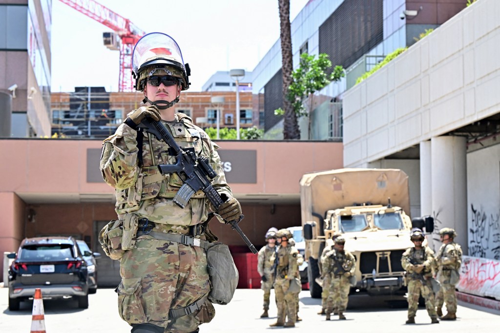 Armed National Guard member with a rifle and riot helmet stands outside a federal building with graffiti.