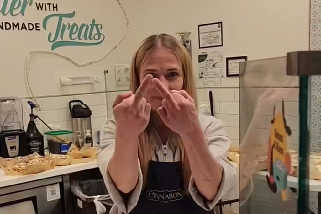 A woman wearing a Cinnabon apron gestures with two middle fingers at the camera.