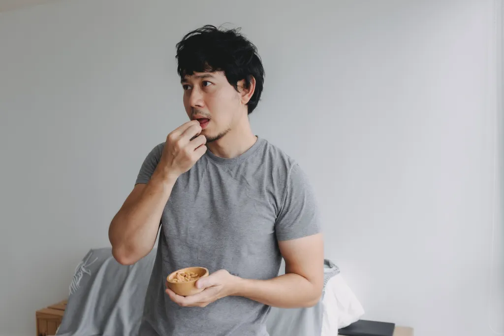 Asian man standing and eating peanuts from a small bowl.