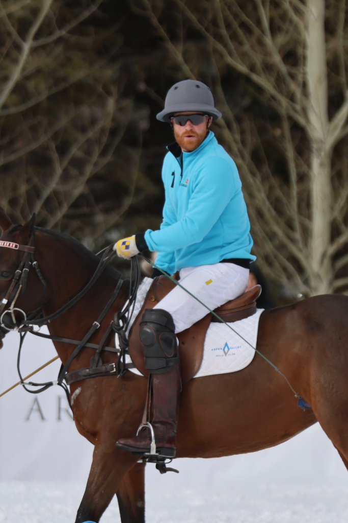 Prince Harry on horseback in a polo match, wearing a gray helmet and blue shirt.