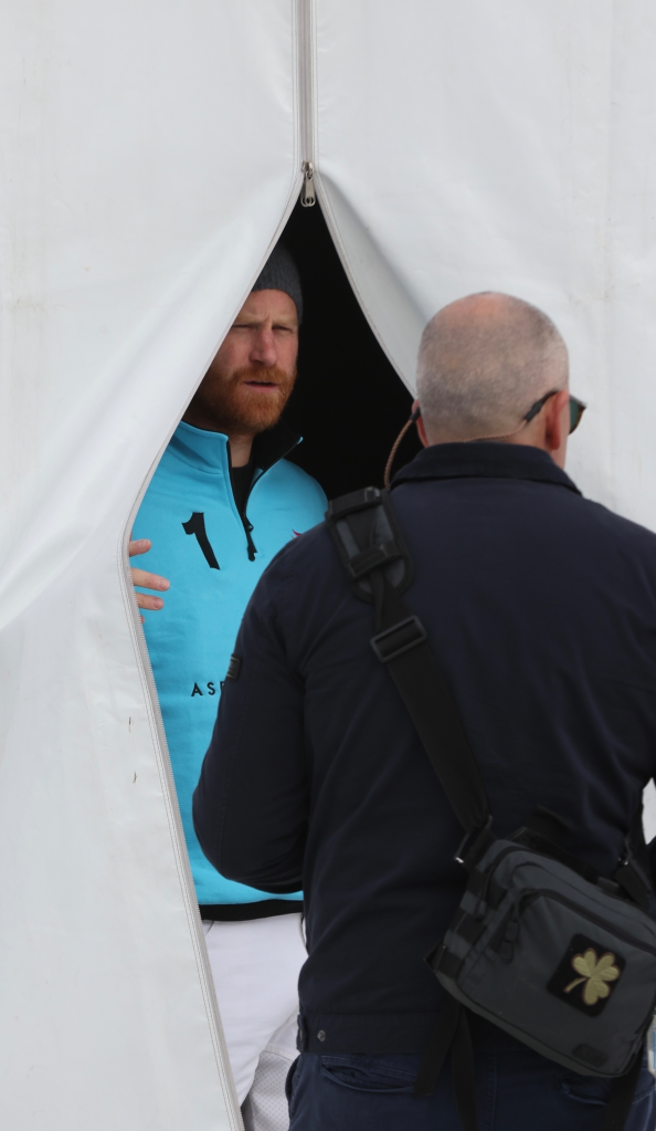 Prince Harry, with a red beard and wearing a light blue polo shirt and grey beanie, looks out from an opening in a white tent, while a bald man in a dark blue jacket and wearing a black shoulder bag stands with his back to the camera.
