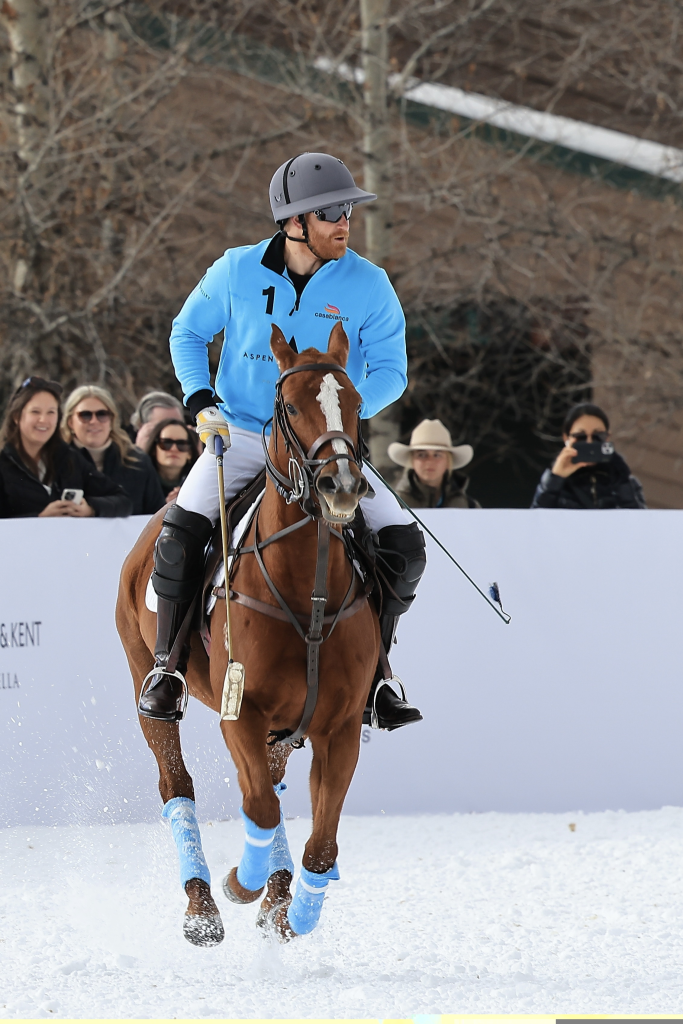 Prince Harry playing polo in Aspen on a brown horse, wearing a blue shirt, white pants, a gray helmet, and sunglasses.