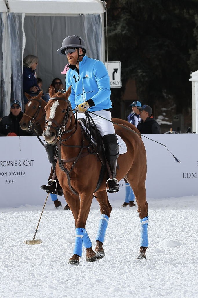 Prince Harry playing polo in Aspen, dressed in a light blue jersey, white pants, and a helmet, riding a brown horse with blue leg wraps on a snow-covered field.