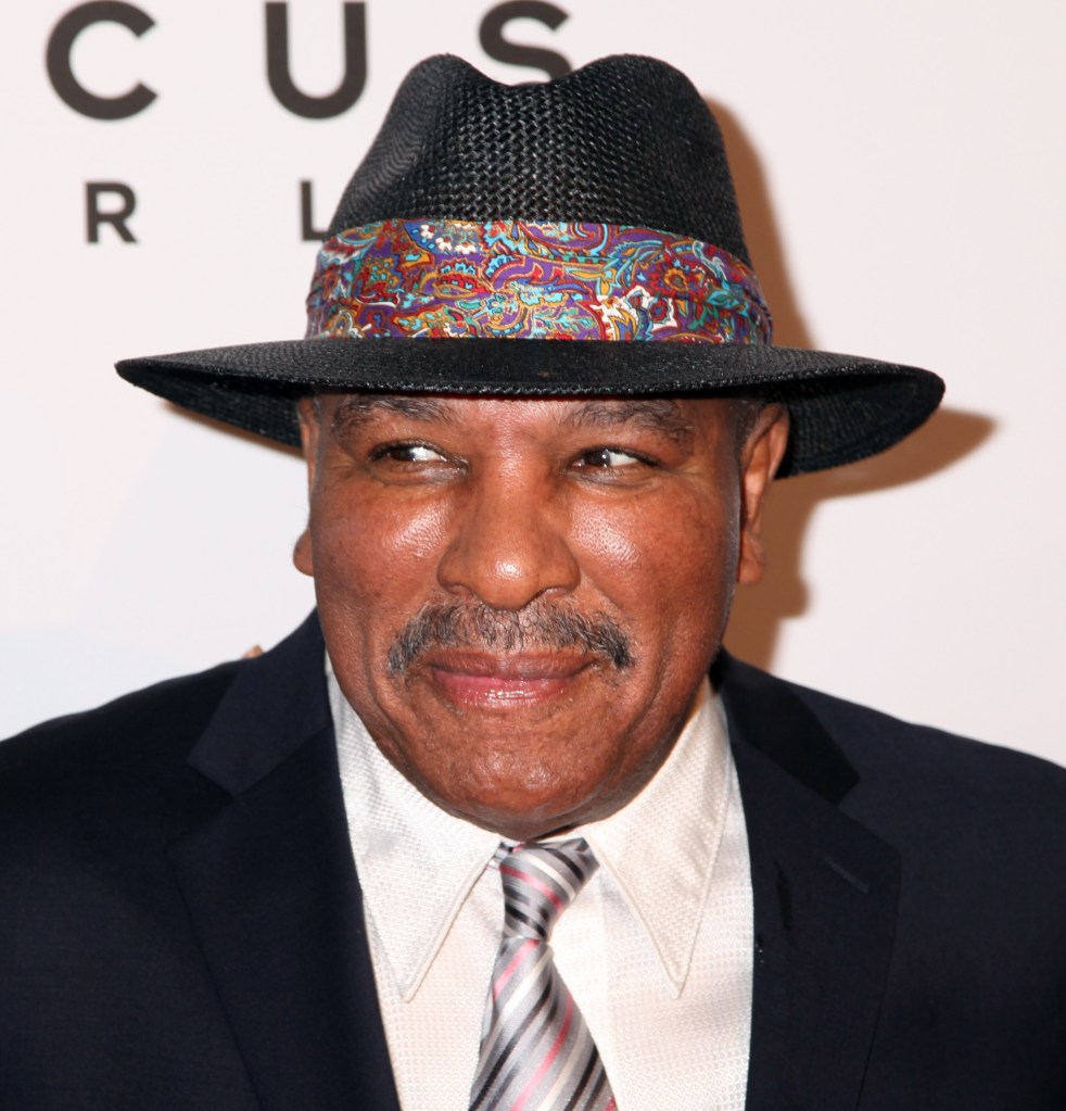 Rahaman Ali, wearing a black fedora with a colorful band, a black suit jacket, and a striped tie.