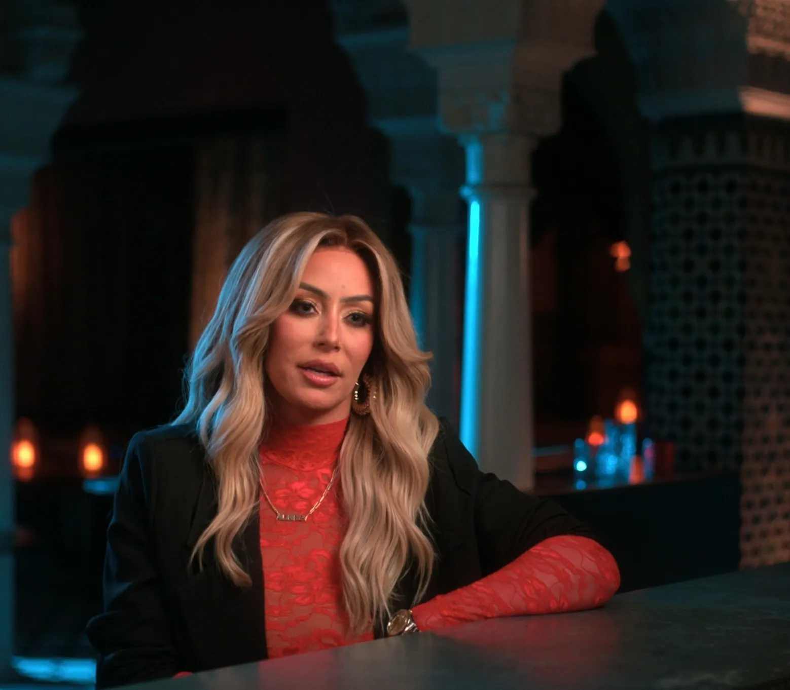 Aubrey O'Day in a red lace top and black jacket, leaning on a dark surface.