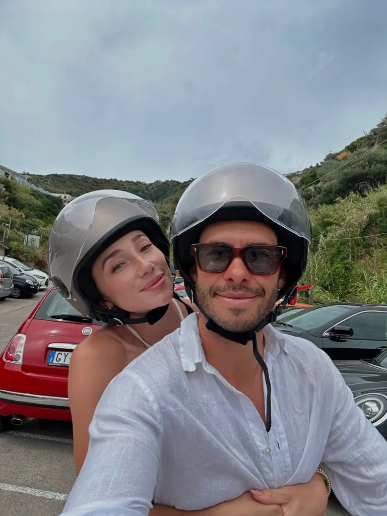 Aurora Culpo and Zac in helmets, smiling, with hills and cars in the background.