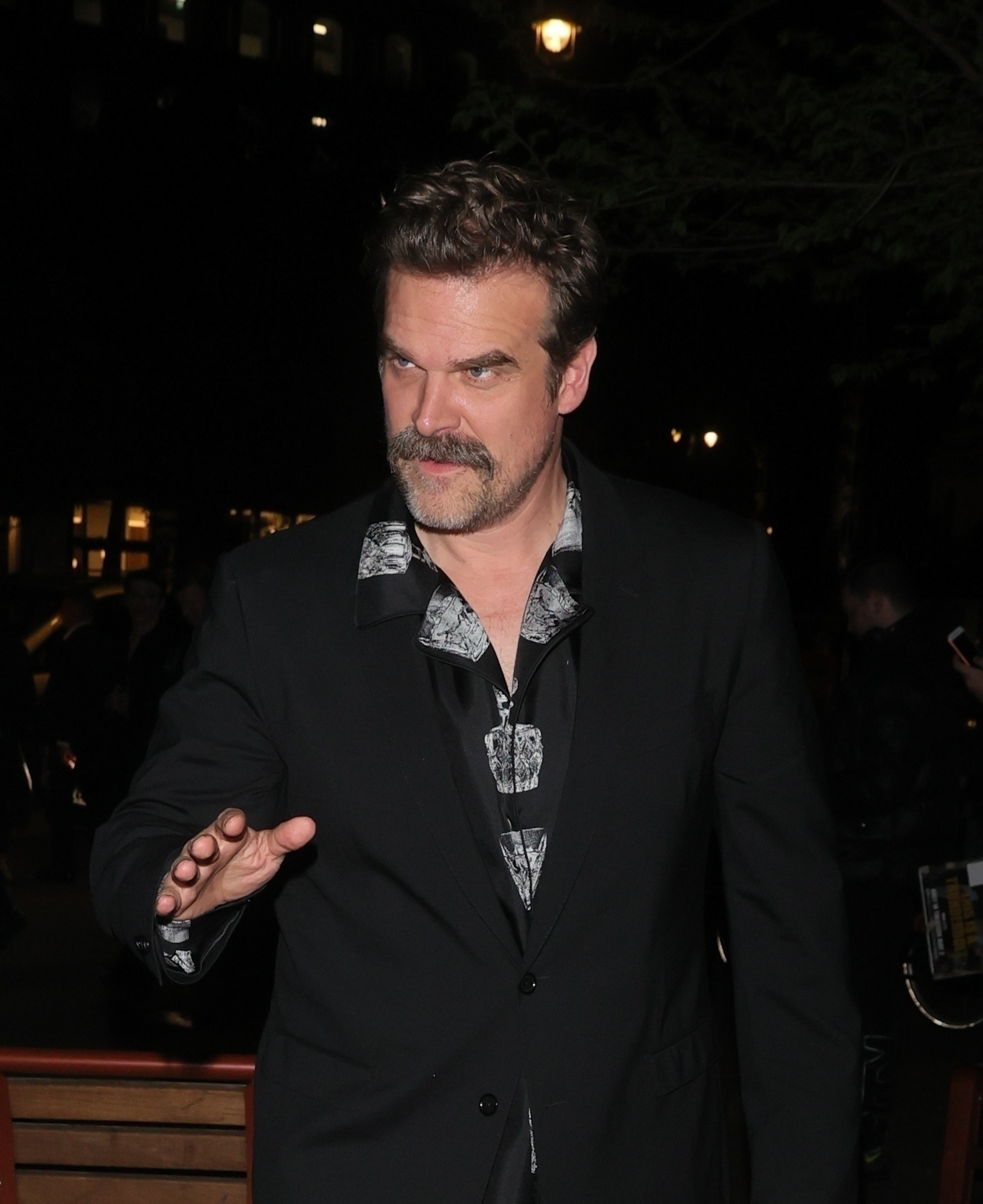 David Harbour in a black blazer and patterned shirt with his hand raised.